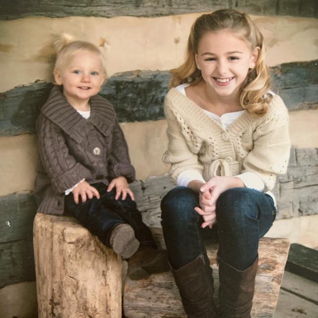 Chloe Lukasiak in a cream color cardigan and noir jeans smiling with her sister in a grey cardigan and black jeans.